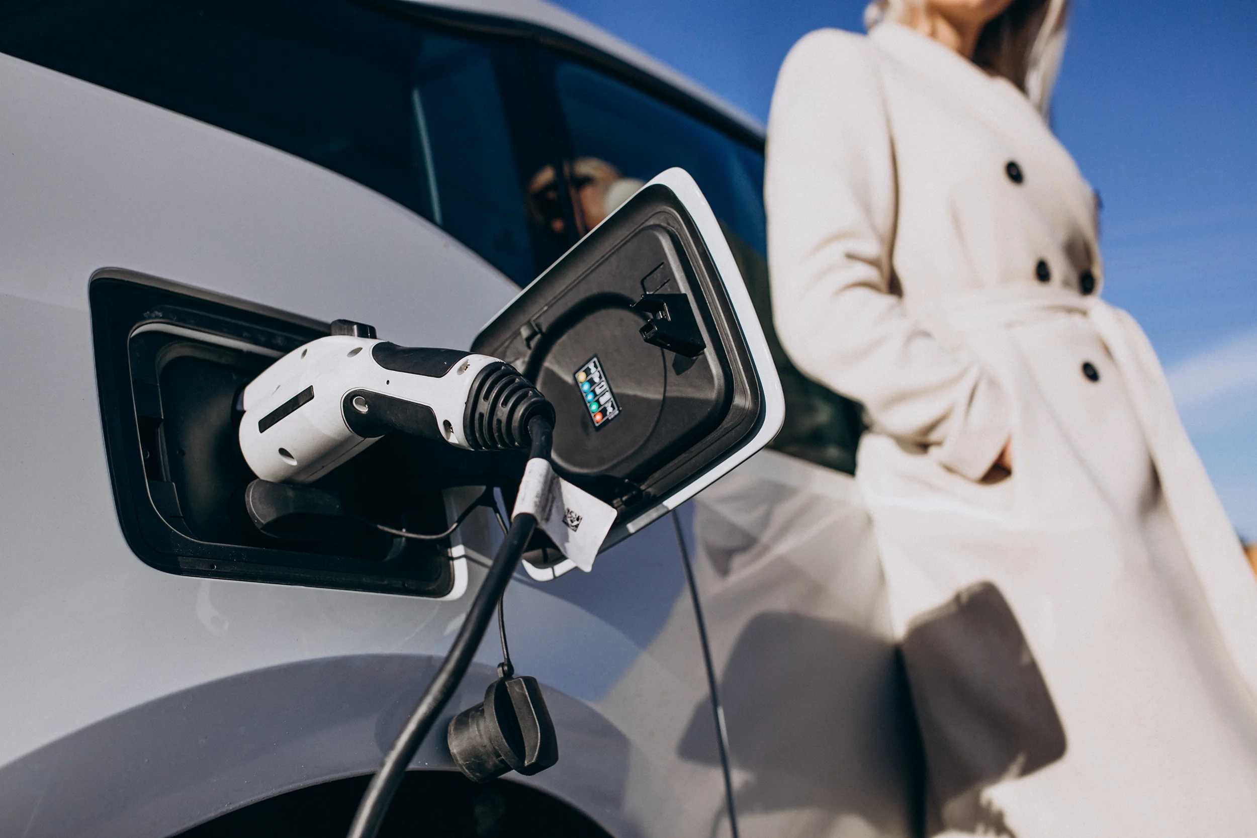 A woman in a cream coat standing beside a silver electric car as it charges, the connector plugged into the car's charging port under a clear blue sky.